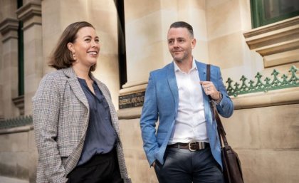 UQ MBA students Gabrielle Woodbridge and Andrew Ward. A woman and a man in business wear walking past a sandstone building